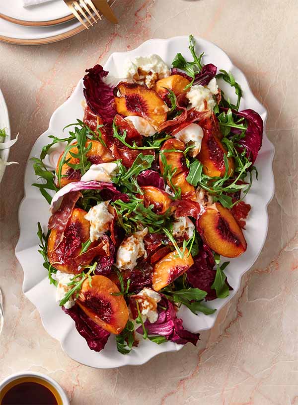 A platter with a salad. The salad is made up of air-fried peaches, prosciutto, leafy greens and buffallo mozzarella.
