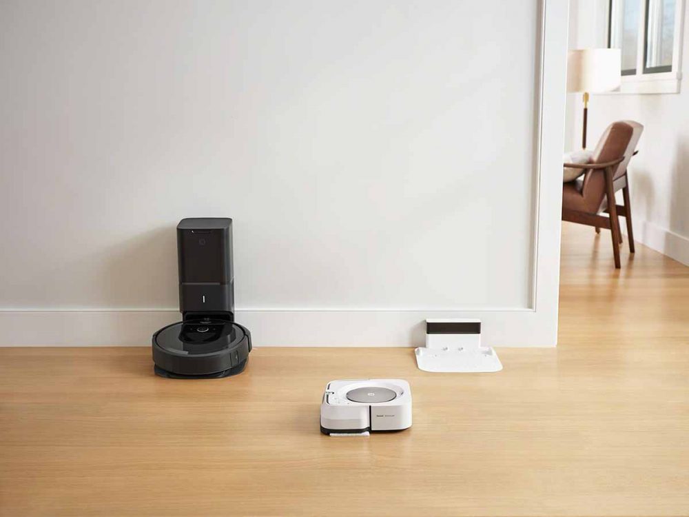 The iRobot Braava jet m6 Robot Mop cleaning the floors while the iRobot Roomba i7+ Robotic Vacuum sits in the dock.
