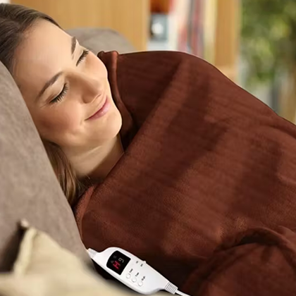 Woman relaxing under a brown electric blanket with digital controller on a couch