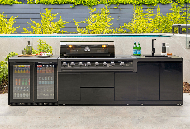 Black outdoor kitchen with built-in BBQ, double drinks fridge, sink, storage cabinets and stone-look benchtop. 