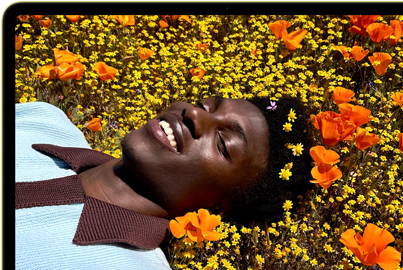 MacBook Neo, citrus colour, screen shows a smiling person lying in a sunny field of flowers